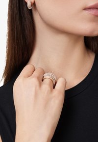 Anelli tono oro con più file di piccole pietre, indossati su una mano. Lo sfondo presenta una spalla e un collo con una maglietta nera.