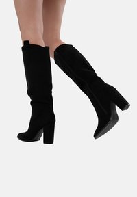 Botas altas de ante negro hasta la rodilla con punta cuadrada, tacón grueso y detalle de cremallera en el interior. Textura suave con una silueta ajustada.