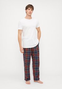 T-shirt blanc à manches courtes associé à un pantalon à carreaux bleus avec des lignes rouges et vertes. Le modèle est pieds nus avec les mains dans les poches.