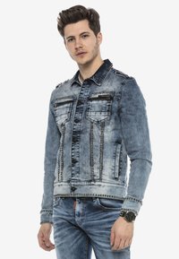 Denimjack in vervaagd blauw met metalen studs langs de naden en zakken. Bevat twee borstzakken en een knopenfrontsluiting.
