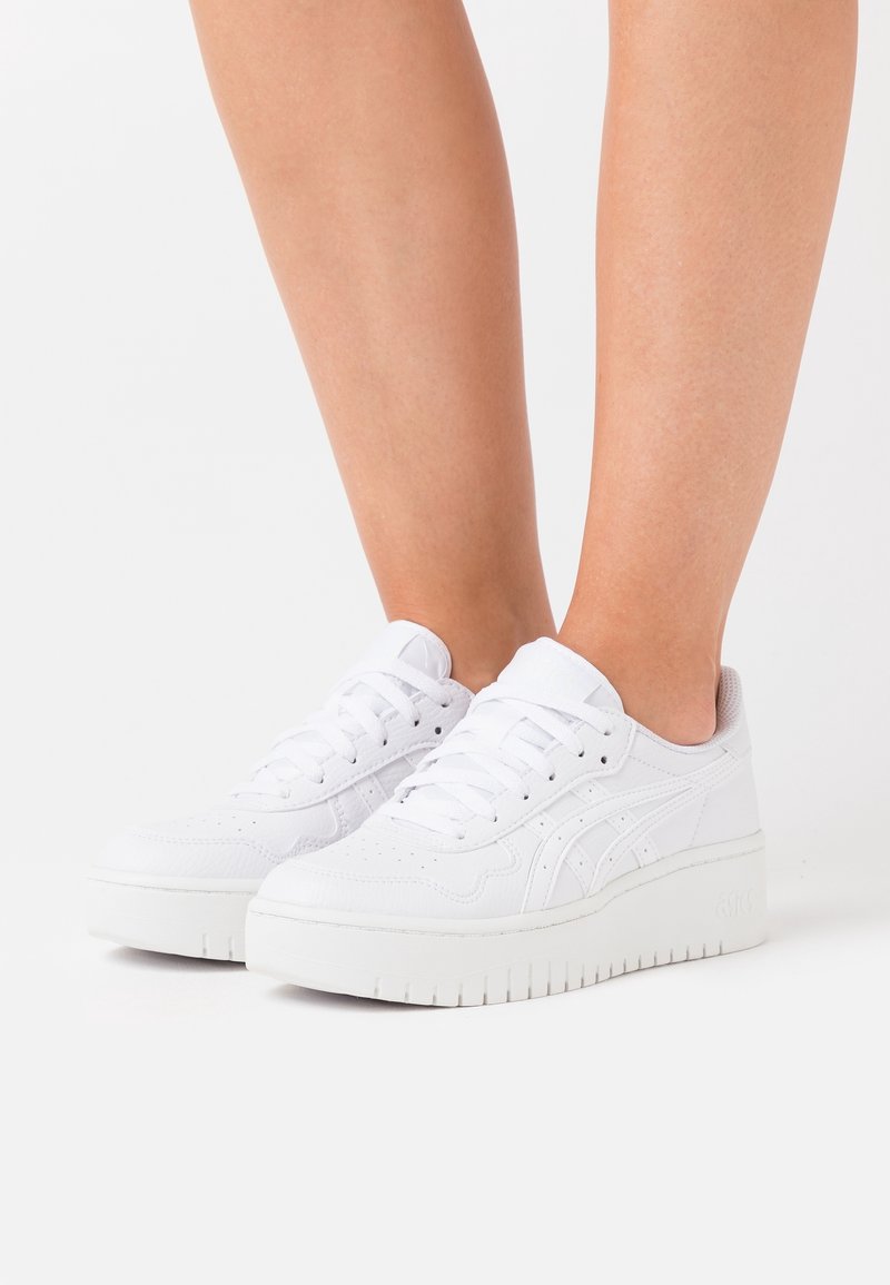 Nogi w białych sneakersach na platformie ze sznurowadłami, stojące na białej powierzchni na jednolitym tle.