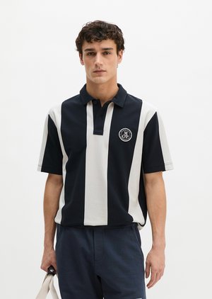 Junger Mann mit lockigem Haar trägt ein marineblau-weiß vertikal gestreiftes Polo mit Logo und hält einen beigen Taschenhenkel an seiner Seite.