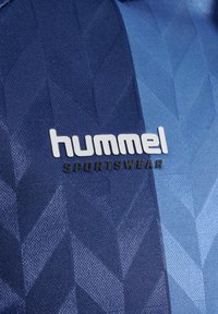 Tumši zils sporta krekls ar teksturētu ģeometrisku rakstu. Uz tā redzams zīmola nosaukums "hummel" baltā krāsā un "SPORTSWEAR" melnā krāsā zem tā.