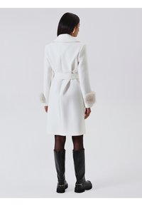 Weißer Mantel mit einem Gürtel, der Fellakzente an den Ärmelbündchen aufweist. Lange Ärmel, gerader Schnitt und klassischer Kragen. Schwarze kniehohe Stiefel.