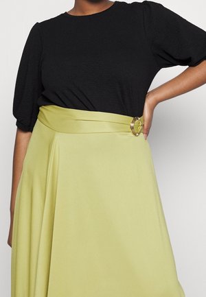 Femme portant un haut noir à manches bouffantes rentré dans une jupe taille haute vert clair avec une large ceinture et une boucle circulaire dorée.