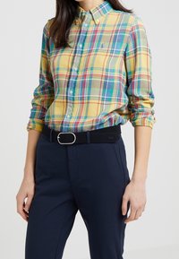 Camicia a quadri gialla, verde e blu, con colletto button-down. Presenta maniche lunghe, vestibilità slim e un piccolo dettaglio del logo. Indossata con una cintura nera.