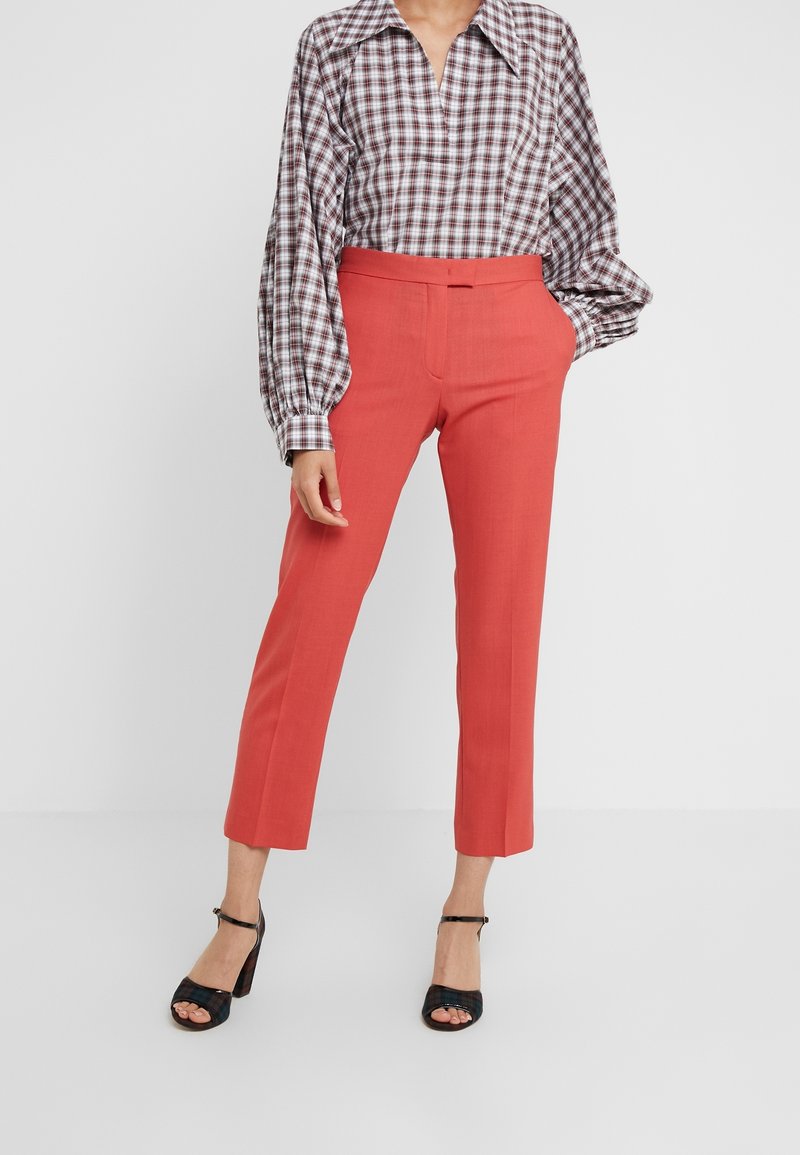 Rode op maat gemaakte broeken met een gladde textuur, gecombineerd met een geruit blouse met een kraag en volumineuze mouwen, afgewerkt met zwarte hakken.