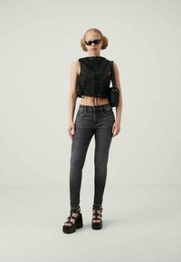 Νέος/νέα που φορά μαύρη μανικέτα χωρίς μανίκια, σκούρα στενά τζιν, πλατφόρμες, μαύρα γυαλιά ηλίου και κρατά μια μαύρη τσάντα ώμου.