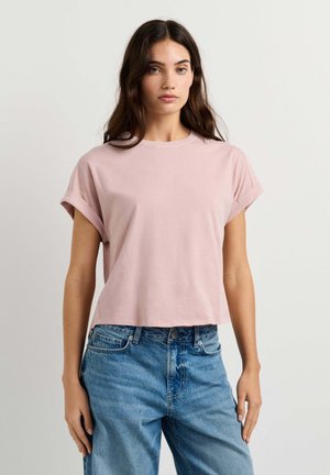 Jonge vrouw met lang bruin haar die een lichtroze T-shirt met korte mouwen en een blauwe high-waisted jeans draagt tegen een effen witte achtergrond.