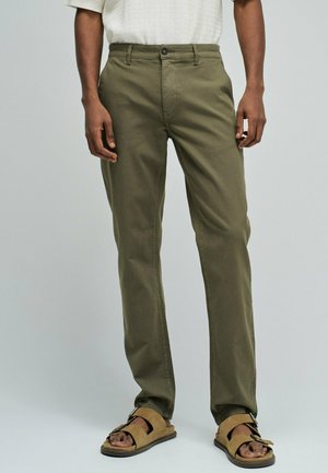 Homme portant un pantalon droit vert olive et des sandales à boucle marron clair, debout devant un fond uni gris clair.
