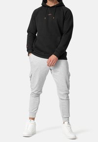 Henkilö, jolla on päällään musta huppari, vaaleanharmaat cargo-housut ja valkoiset lenkkarit, seisoo kädet taskuissa yksinkertaista taustaa vasten.