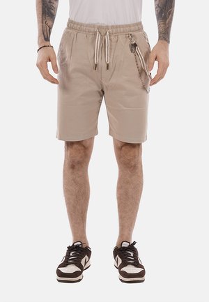 Uomo che indossa pantaloncini beige con coulisse, tasca laterale con zip e un accessorio a catena, e sneakers nere e bianche.
