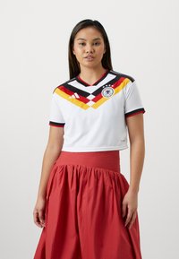 Jonge vrouw die een wit Duits voetbalshirt draagt met rode, zwarte en gele strepen en een lange rode rok, staand tegen een effen achtergrond.