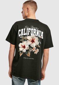 Sort bomulds-T-shirt med et blomstermotiv bestående af hibiscusblomster i hvid og rød samt teksten "Los Angeles CALIFORNIA" i fede hvide bogstaver.