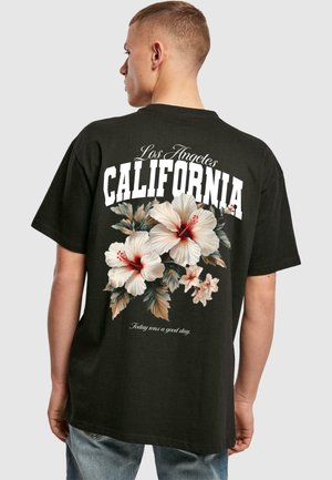 Melna kokvilnas T-krekls ar ziedu dizainu, kurā ir hibiska ziedi baltā un sarkanā krāsā, un teksts "Los Angeles CALIFORNIA" biezajās baltajās burtiņās.