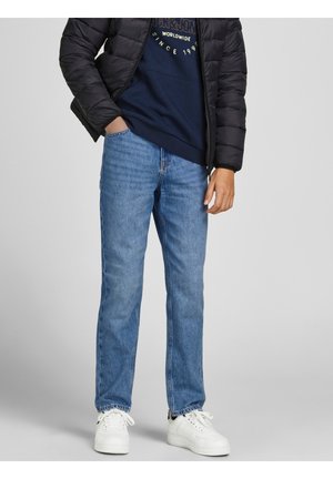 Person iført lyseblå jeans med lige ben, hvide sneakers, marineblå sweatshirt og sort quiltet jakke, der står mod en grå baggrund.
