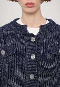 Personne portant un cardigan en maille épaisse bleu marine avec des boutons métalliques et des poches à rabat par-dessus une chemise blanche, montrant les détails du col et des épaules.