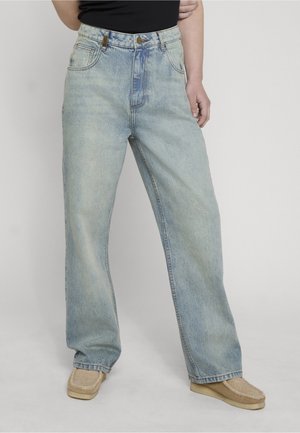 Osoba nosząca jasnoniebieskie szerokie jeansy i beżowe wsuwane buty, stojąca na tle prostego jasnego tła.