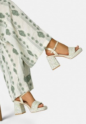 Woman's feet wearing beige block-heeled sandals with ankle straps, paired with loose white pants featuring green floral patterns.