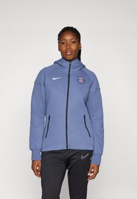 Blå hettejakke med høy krage, svarte glidelåsdetaljer og Paris Saint-Germain-logo på brystet, laget av elastisk stoff.