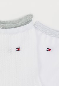 Άσπρες κάλτσες με ρίγες, διαθέτουν ασημί μεταλλικά μανσέτες στον αστράγαλο και μικρό κεντημένο λογότυπο σε κόκκινο, ναυτικό μπλε και άσπρο χρώμα σε κάθε πλευρά.