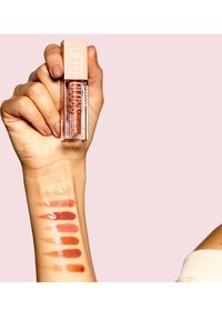 Maybelline Lifter Gloss in clear container with a light pink lid; swatches of various glossy lip colors on a wrist, ranging from nude to deep brown.
