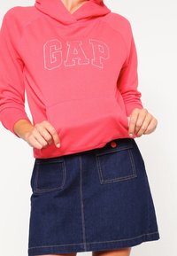 Mikina s kapucí v jasně růžové barvě s logem "GAP", doplněná tmavou džínovou minisukní se dvěma předními kapsami a detaily v podobě oranžových knoflíků.