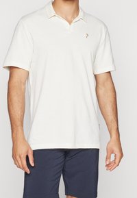 Kortærmet polo-shirt i creme med en foldet krave, der har et lille guld logo på brystet, lavet af blødt bomuldsstof.