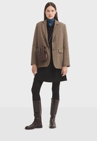 Brązowa marynarka w kratę, czarny golf, czarna sukienka, czarne rajstopy i brązowe skórzane buty za kolano. Mała brązowa torebka na ramieniu.