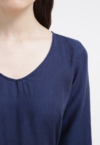 Blusa blu navy con scollatura a V, maniche lunghe e una texture sottile. I dettagli di cucitura lungo la scollatura aggiungono un tocco raffinato.