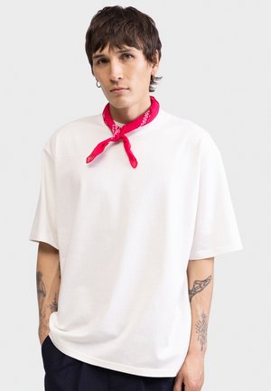 Νεαρός άνδρας με κοντά καστανά μαλλιά, φορώντας λευκό άνετο t-shirt, κόκκινο μαντίλι γύρω από το λαιμό και τατουάζ σε κάθε πήχη.