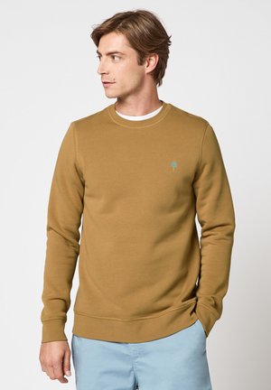 Homme portant un sweat-shirt à manches longues marron moutarde avec un petit logo d'arbre vert, pantalon bleu clair, regardant vers la gauche sur un fond uni.