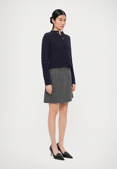 Navy knitted cardigan with cable pattern, five black buttons, and a cropped design, paired with a gray skirt and black heeled shoes.
