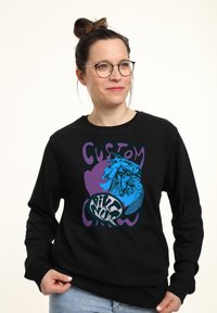 Svart sweatshirt med en färgglad grafik av en motorcykel, med "CUSTOM" i lila och "BIKE NUR" i fet stil. Material av bomullsblandning.