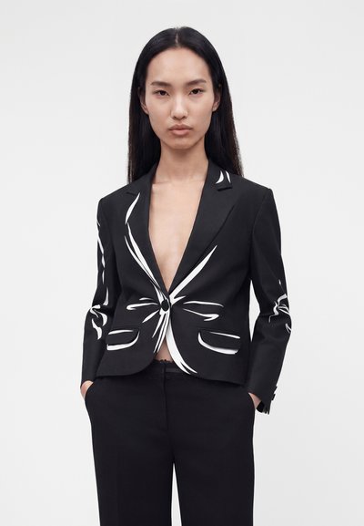 Γυναίκα με μαύρο σακάκι με λευκές αφηρημένες γραμμές, μαύρο παντελόνι, με τα χέρια στις τσέπες, στέκεται μπροστά σε απλό ανοιχτό γκρι φόντο.