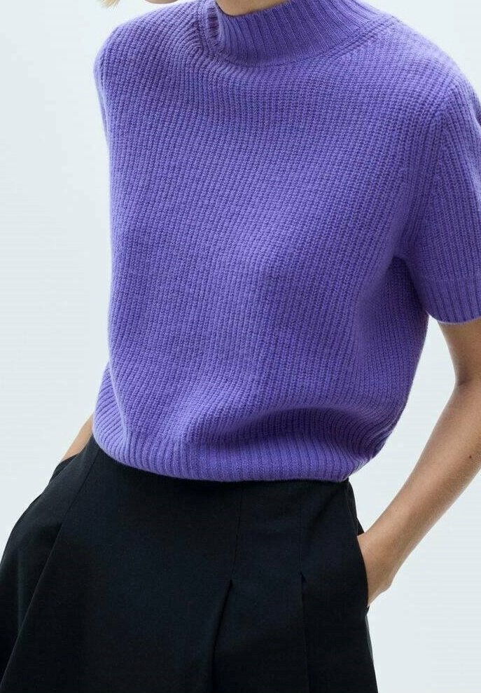 Femme portant un pull violet côtelé à manches courtes rentré dans un pantalon noir taille haute, les mains dans les poches.