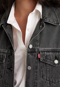 Chaqueta de mezclilla gris con herrajes plateados, presenta un collar puntiagudo y dos bolsillos en el pecho. Lleva puesta una camisa blanca de botones. Etiqueta del logo roja visible.