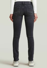Donkergrijze skinny jeans met een gladde textuur, voorzien van zakken aan de achterkant, riemlussen en stiksels langs de benen. Draagt met lichtgekleurde schoenen.