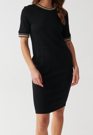 Kobieta ubrana w dopasowaną czarną sukienkę do kolan z krótkimi rękawami i beżowymi paskami na kołnierzyku i mankietach na jednolitym tle.