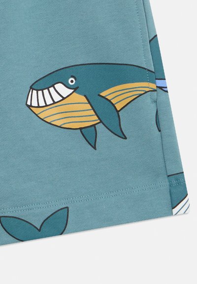 Τυρκουάζ ύφασμα με σχέδιο φάλαινας που έχει λευκό χαμόγελο και κίτρινες ρίγες. Περιλαμβάνει μπλε λεπτομέρειες και διακριτικό μοτίβο ουράς.