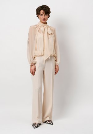 Beige, transparente Bluse mit Bindekragen und gerafftem Bund. Kombiniert mit hochtaillierten, cremefarbenen, weit geschnittenen Hosen. Auffällige schwarz-weiße gemusterte Schuhe.