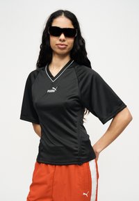 Γυναίκα με μακριά σκούρα μαλλιά, που φοράει μαύρο αθλητικό μπλουζάκι Puma, κόκκινα σορτς Puma και μεγάλα μαύρα γυαλιά ηλίου, μπροστά σε απλό φόντο.