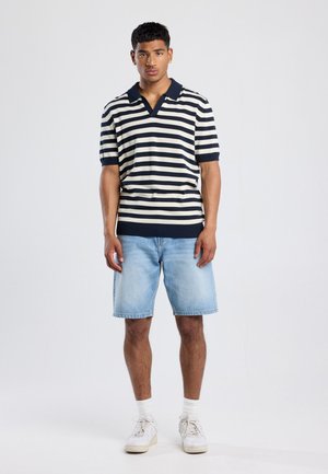 Marineblauw en crème gestreepte polo met korte mouwen, V-hals en geribde zoom, gecombineerd met lichte denimshorts en witte sneakers.