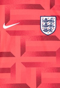 Camiseta roja de Nike con un patrón geométrico y acentos en rojo oscuro, y un escudo con tres leones y detalles florales en el lado izquierdo del pecho.