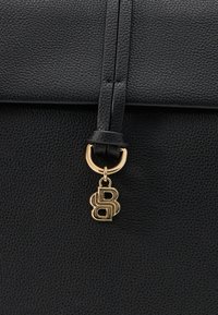 Μαύρη δερμάτινη τσάντα με υφή, με χρυσό μεταλλικό κρίκο και ένα κρεμαστό χρυσό λογότυπο προσαρτημένο σε δερμάτινο λουρί.