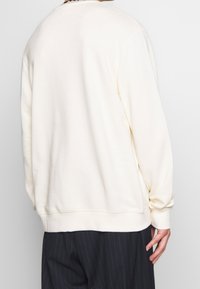 Felpa di colore crema con una vestibilità rilassata, polsini e orlo a coste, texture morbida, design semplice e senza loghi o motivi visibili.