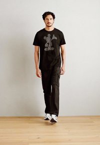 Camiseta de algodón negra con un diseño gráfico, combinada con pantalones cargo negros. El modelo lleva zapatillas deportivas plateadas. Fondo minimalista.