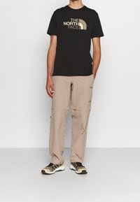 Black t-shirt with a gold logo, paired with light beige cargo pants and sneakers with a black and brown design. Simple, functional outdoor wear.
