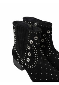 Bottines en suède noir avec des embellissements métalliques en forme d'engrenage et des clous en argent, talon bas, et fermeture éclair sur le côté.