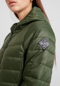 Grüne Puffjacke mit gestepptem Design, Kapuze und schwarchem Hollister-Logo-Patch auf der linken Schulter. Glatte, glänzende Textur.